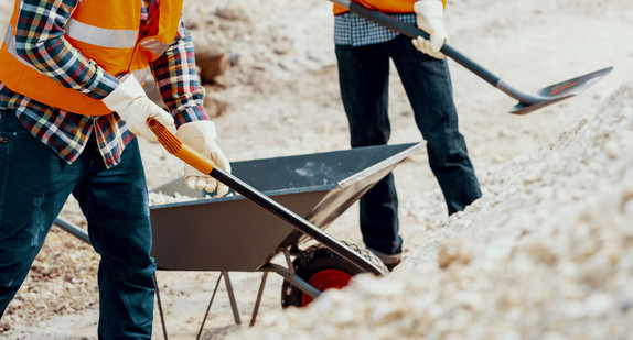 Bauarbeiter arbeiten mit Schaufeln und Schubkarren an einer Baustelle.