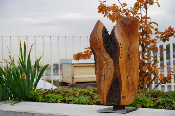 Holz-Skulptur auf Dachterrasse. Im Hintergrund sind Bienenkästen zu sehen.