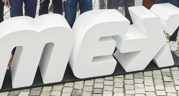Um große Holzbuchstaben mit dem Titel MEX stehen mehrere Menschen zu einem Gruppenbild zusammen.