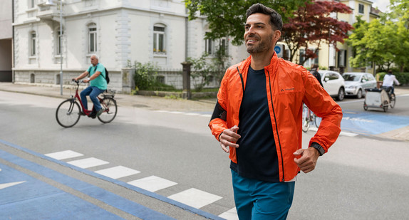 Ein Mann joggt entspannt und gut gelaunt über eine Straßenkreuzung in einem Wohngebiet. Parallel zu ihm fahren Radfahrerinnen und Radfahrer an ihm vorbei.