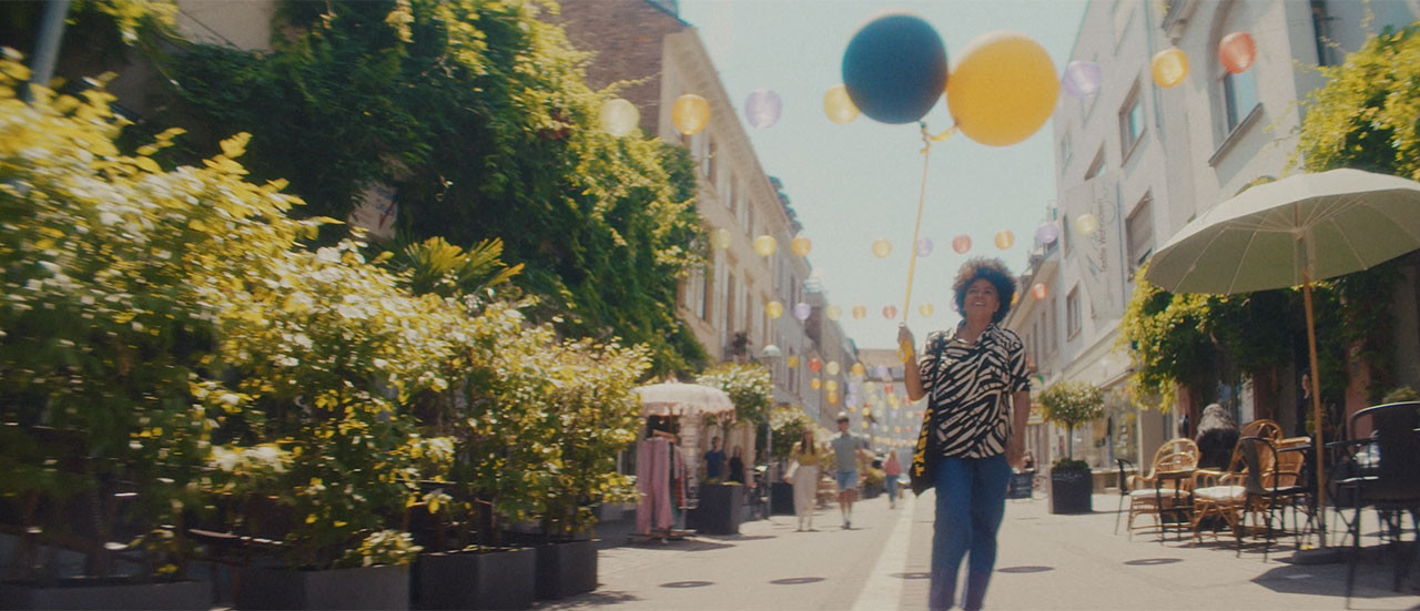 Eine Frau spatziert lachend in einer einladenden Fußgängerzone. Sie hält zwei Luftballons in der Hand.