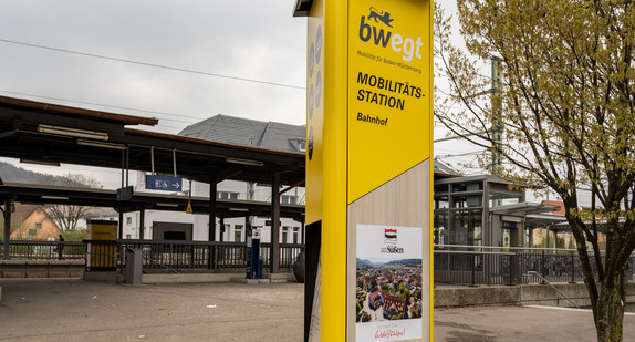 Eine große gelbe Informationssäule steht auf einem Bahnhofsvorplatz.