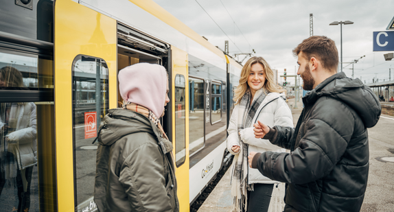 Drei junge Menschen an einem Bahnsteig, vor ihnen Schienenfahrzeug mit geöffneten Türen.