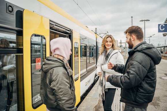 Drei junge Menschen an einem Bahnsteig, vor ihnen Schienenfahrzeug mit geöffneten Türen.