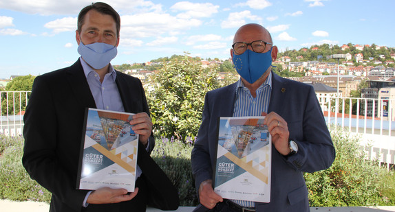 Verkehrsminister Winfried Hermann und Prof. Dr. Tobias Bernecker, beide mit Mundschutz, stehen auf der Terrasse des Verkehrsministeriums und halten beide ein Heft mit der Aufschrift "Güterverkehrskonzept" in der Hand.