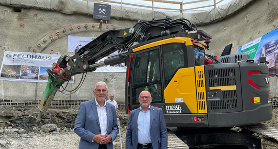 Landrat Helmut Riegger und Verkehrsminister Winfried Hermann stehen vor einem großen Bagger. Im Hintergrund sieht man den eingezeichneten Tunnel der Hermann-Hesse-Bahn.