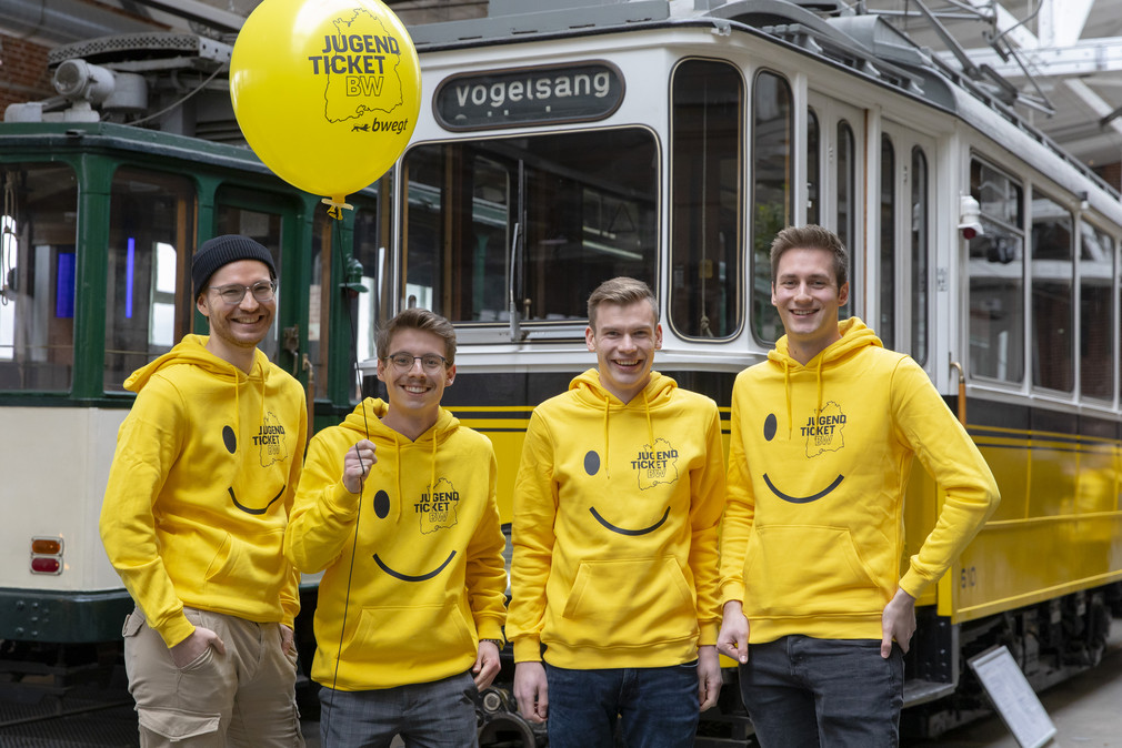 Vier fröhliche junge Männer in gelben Hoodies mit JugendticketBW-Design und mit einem ebensolchen Luftballon vor einer alten Straßenbahn.