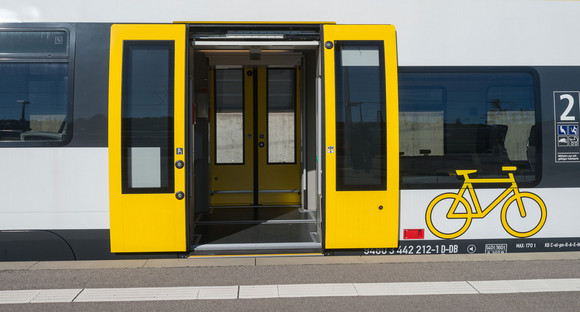Ein weiß-gelber bwegt-Zug steht mit offener Türe an einem Bahnsteig.