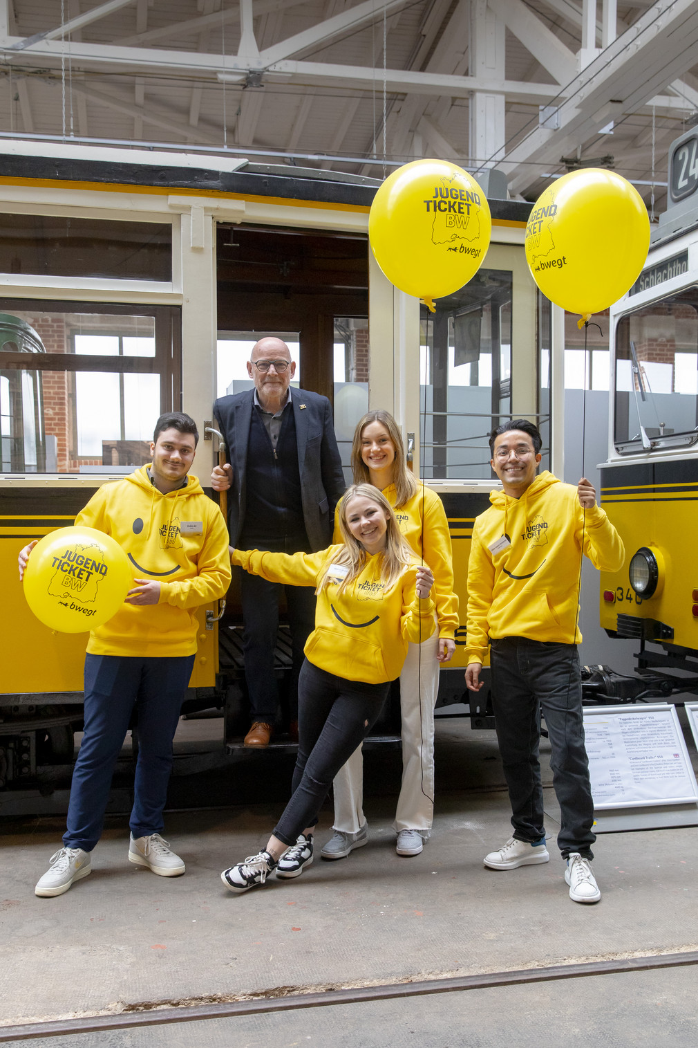 Vier fröhliche junge Menschen in gelben Hoodies mit JugendticketBW-Design und mit drei ebensolchen Luftballons vor einer alten Straßenbahn, während Minister Hermann im Eingangsbereich der Straßenbahn steht.