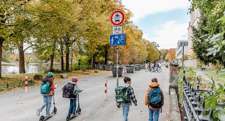 Kinder gehen zu Fuß und mit dem Tretroller zur Schule. Ein Schild zeigt den verkehrssicherten Schulweg an. 