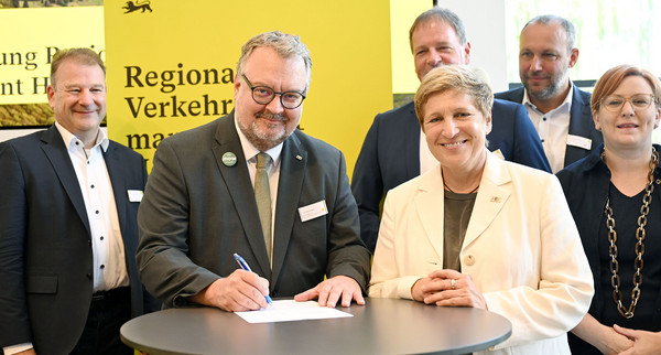 Ein Mann und eine Frau stehen nebeneinander. Der Mann unterzeichnet einen Vertrag. Beide lächeln in die Kamera.