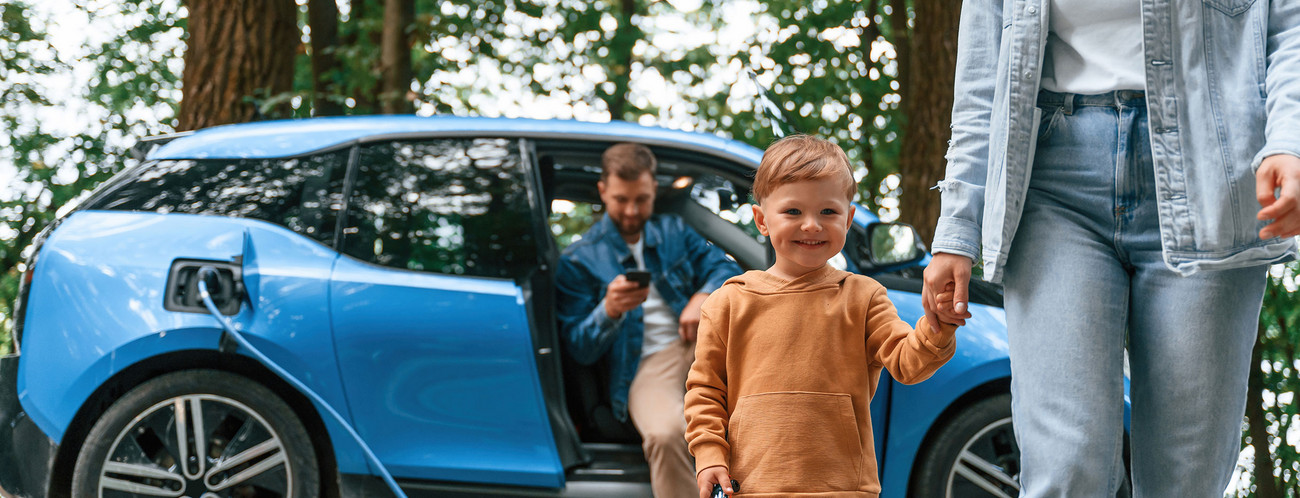 Ein Kind und eine Mutter laufen von einem E-Auto weg in dem noch der Vater sitzt.