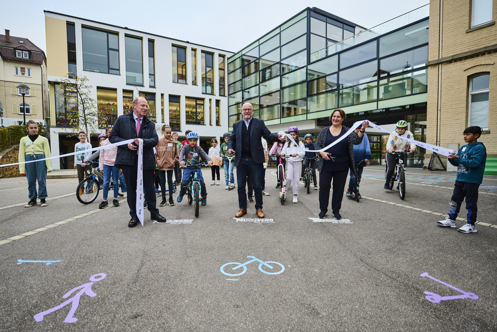Minister Hermann, Ministerin Schopper und Innenstaatssekretär Klenk beim Startschuss von MOVERS, während sie das Band durchschneiden. Im Hintergrund Kinder zu Fuß, mit dem Fahrrad oder Roller.