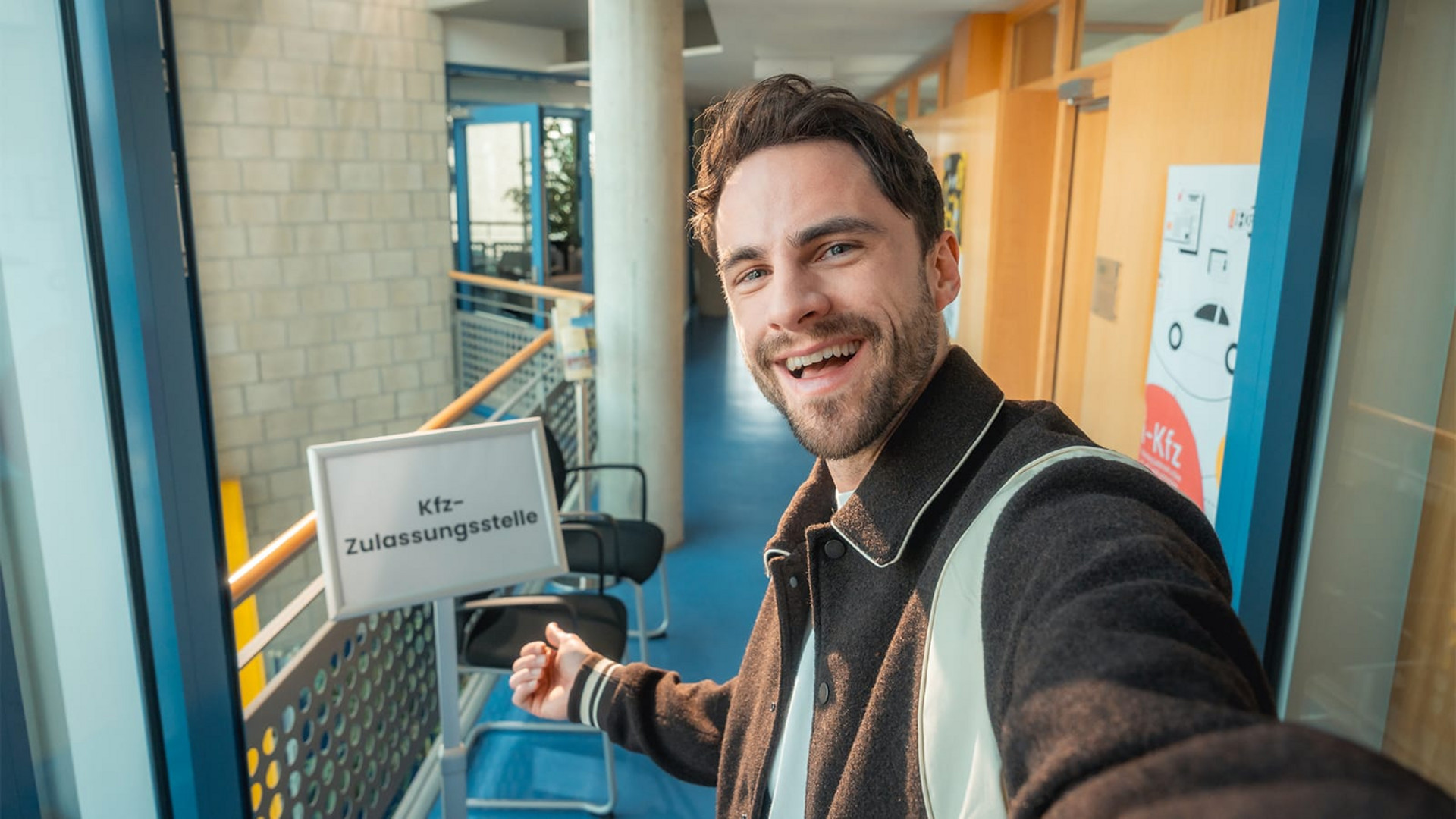 Ein Mann steht in einem langen Flur, lacht freundlich in die Kamera und zeigt auf ein Schild, auf dem Kfz-Zulassungsstelle steht. Das Bild wirkt wie mit der Selfie-Kamera eines Smartphones aufgenommen. 