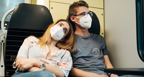 Eine junge Frau und ein junger Mann sitzen zährtlich nebeneinander in einem Zug und tragen eine FFP2-Maske.