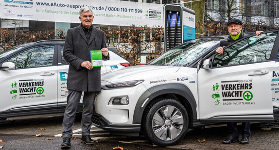 Burkhardt Metzger, Präsident der Landesverkehrswacht Baden-Württemberg, und Verkehrsminister Winfried Hermann stehen auf einem Parkplatz neben einem weißen Elektroauto.