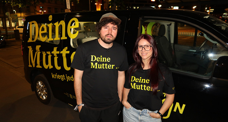 Ein Mann mit Cap und eine Frau stehen vor einem Auto mit der Aufschrift DEINE MUTTER und haben schwarze T-Shirts an mit gelber Schrift &amp;quot;Deine Mutter...&amp;quot;