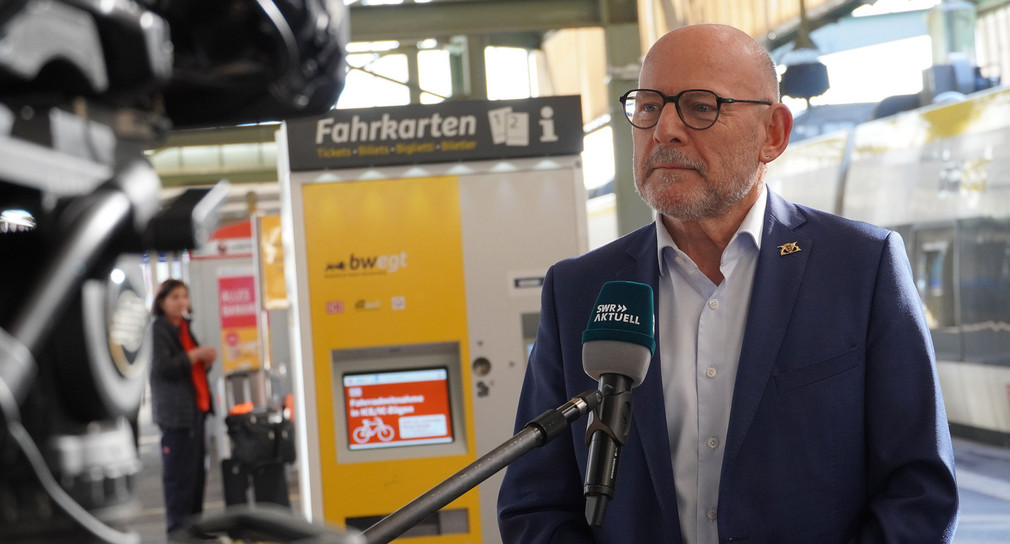 Verkehrsminister Hermann steht vor einer Fernsehkamera an einem Bahnsteig und spricht in ein Mikrofon von SWR Aktuell.