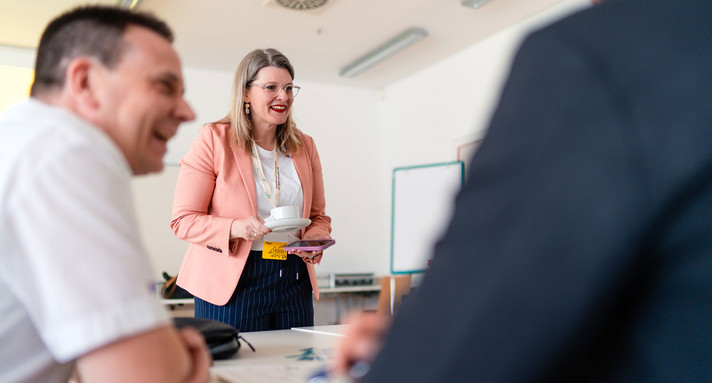 Eine Frau steht an einem Tisch an dem gerade andere eine Gruppenarbeit durchführen.