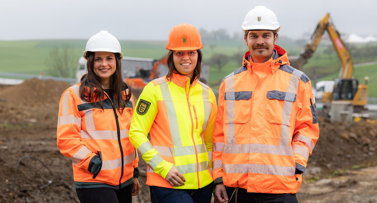 Drei Personen in Sicherheitsausrüstung stehen auf einer Baustelle mit Bagger im Hintergrund.
