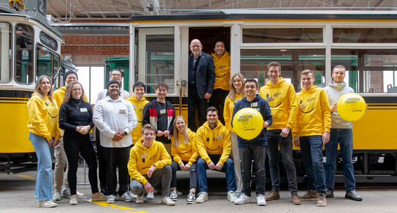 Minister Hermann im Eingangsbereich einer alten Straßenbahn, vor ihm eine Gruppe junger Menschen, teils mit gelben Hoodies im JugendticketBW-Design sowie mit ebensolchen Luftballons.