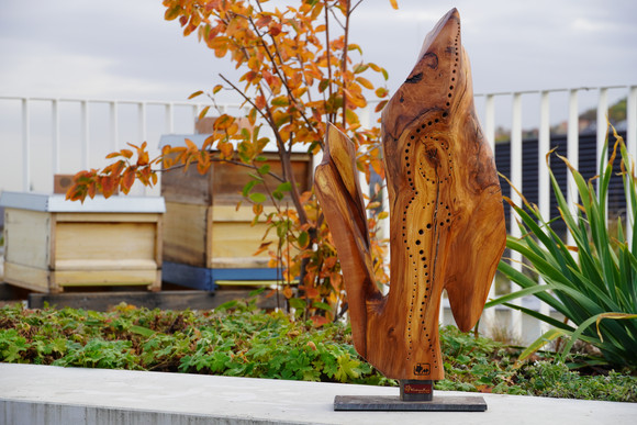 Holz-Skulptur auf Dachterrasse. Im Hintergrund sind Bienenkästen zu sehen.