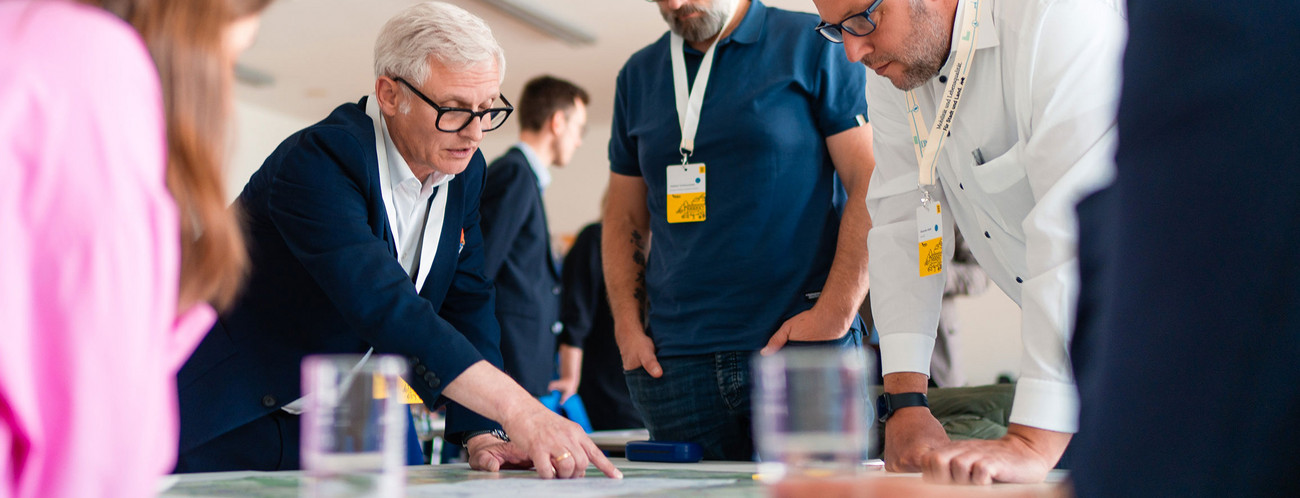Eine Gruppe von Menschen beugt sich über einen Tisch auf dem eine Karte liegt. Ein Mann zeigt auf eine Stelle.
