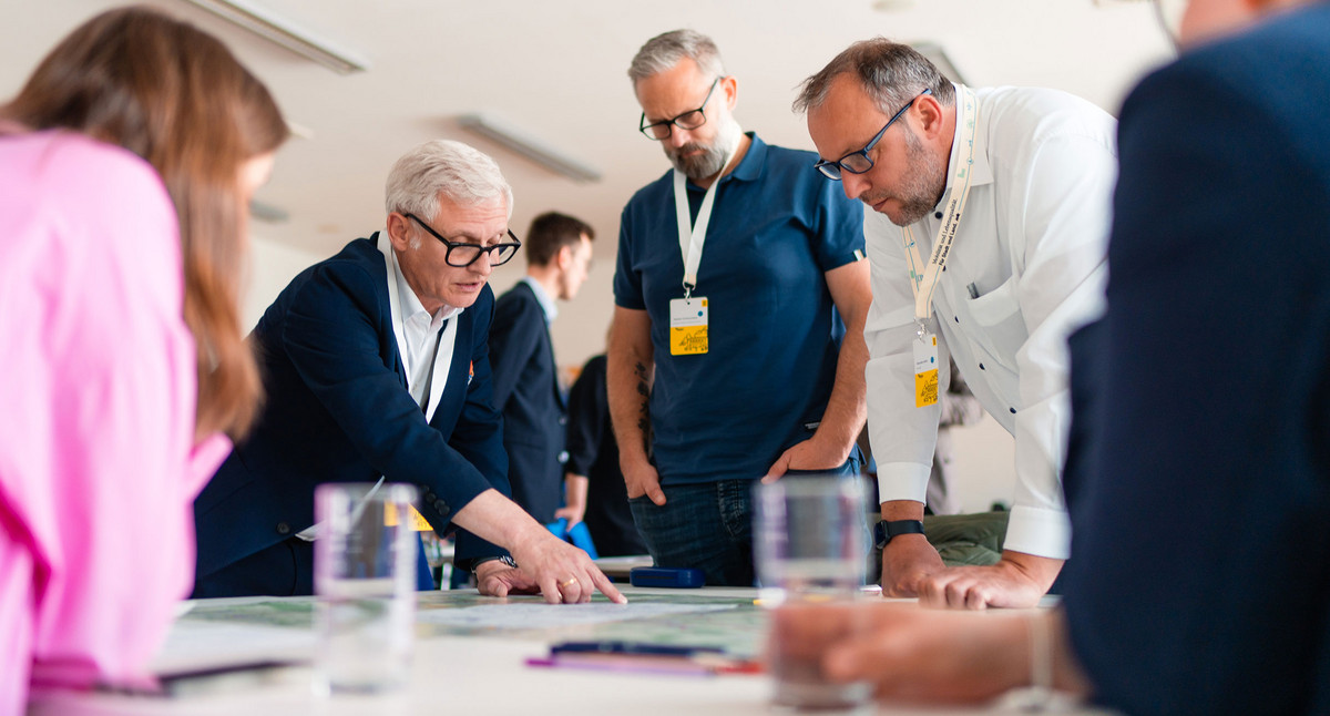 Eine Gruppe von Menschen beugt sich über einen Tisch auf dem eine Karte liegt. Ein Mann zeigt auf eine Stelle.