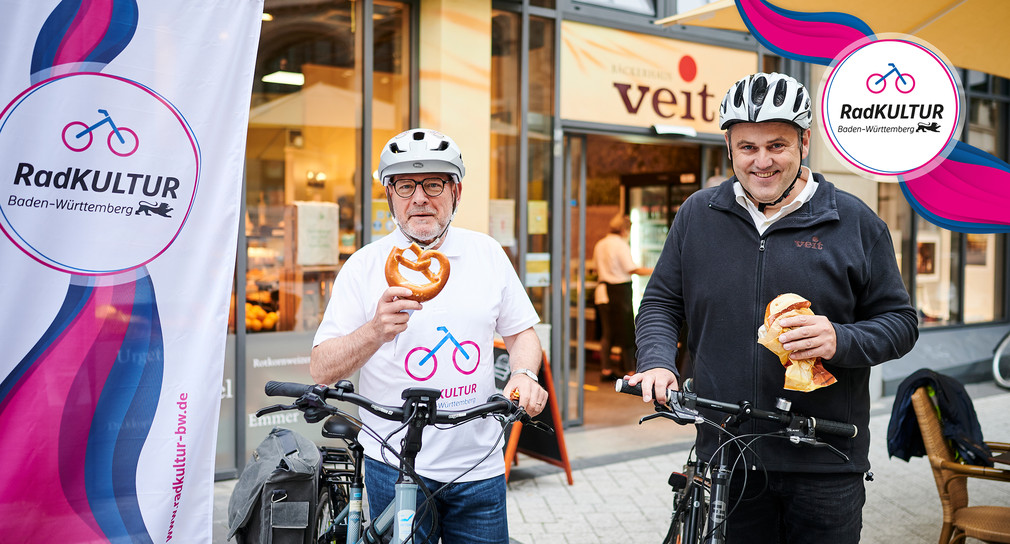 Verkehrsminister Winfried Hermann steht an seinem Fahrrad und präsentiert eine Gratis-Brezel in die Kamera.