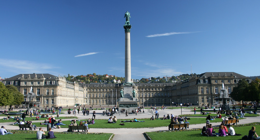 Stuttgarter Schlossplatz im Sommer. Im Zentrum des Platzes steht eine große Säule. Um die Säule liegen auf der Wiese viele Menschen.
