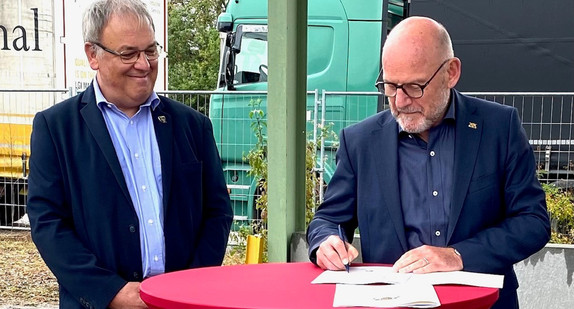 Minister Hermann unterzeichnet die Förderbescheide, neben ihm steht ein anderer Mann