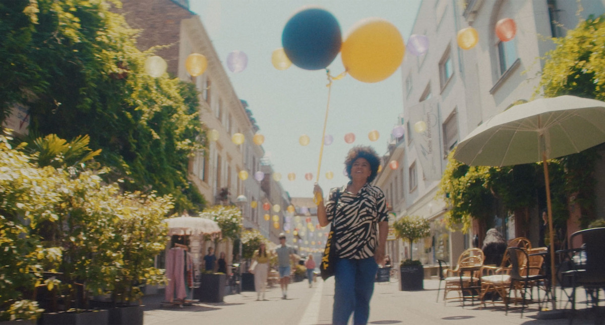 Eine Frau hält zwei große Luftballons in der Hand. Sie spatziert durch eine bunte Fußgängerzone mit vielen grünen Bäumen.