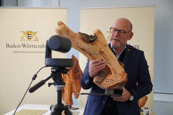 Minister Hermann steht vor einer Kamera und hält eine Holz-Skulptur in den Händen.