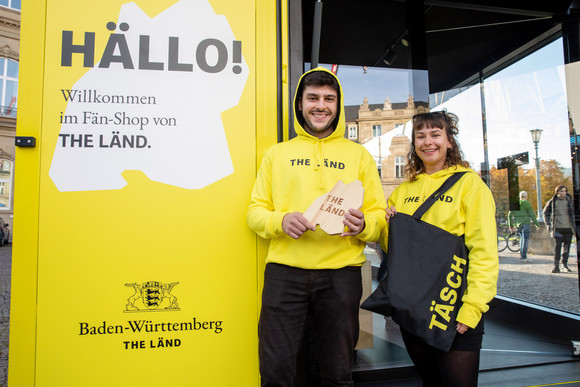 Imagekampagne Baden-Württemberg ist „THE LÄND“ Ein Mann und eine Frau im mobilen „Fän“-Shop in Form eines umgebauten Frachtcontainers vor dem Neuen Schloss in Stuttgart