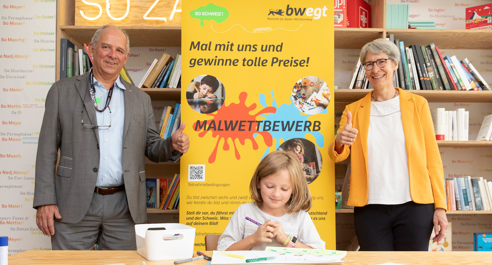 Ein kleines Mädchen sitzt an einem Holztisch und malt ein Bild. Neben ihr stehen Ernst Steinmann (links), Generalkonsul der Schweiz, und Elke Zimmer (rechts), Staatssekretärin im Ministerium für Verkehr, und zeigen einen Daumen hoch in die Kamera.