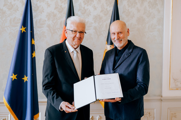 Ministerpräsident Winfried Kretschmann (links) und Tadeusz Matacz (rechts) stehen vor Fahnen und präsentieren die Verleihungsurkunde.