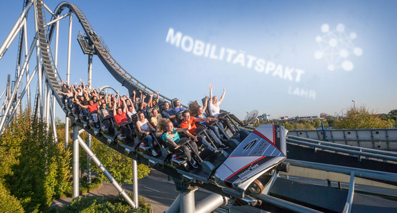 Achterbahn fährt einen Hang hinunter. Im blauen Himmel bilden Wolken den Schriftzug Mobilitätspakt Lahr.