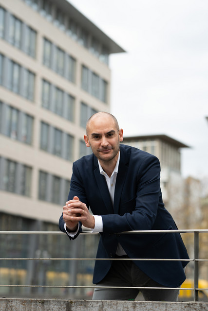 Porträtbilder Minister Dr. Danyal Bayaz Finanzminister Danyal Bayaz lehnt an einem Geländer, im Hintergrund sind moderne Bürogebäude am kleinen Schlossplatz in Stuttgart zu sehen.