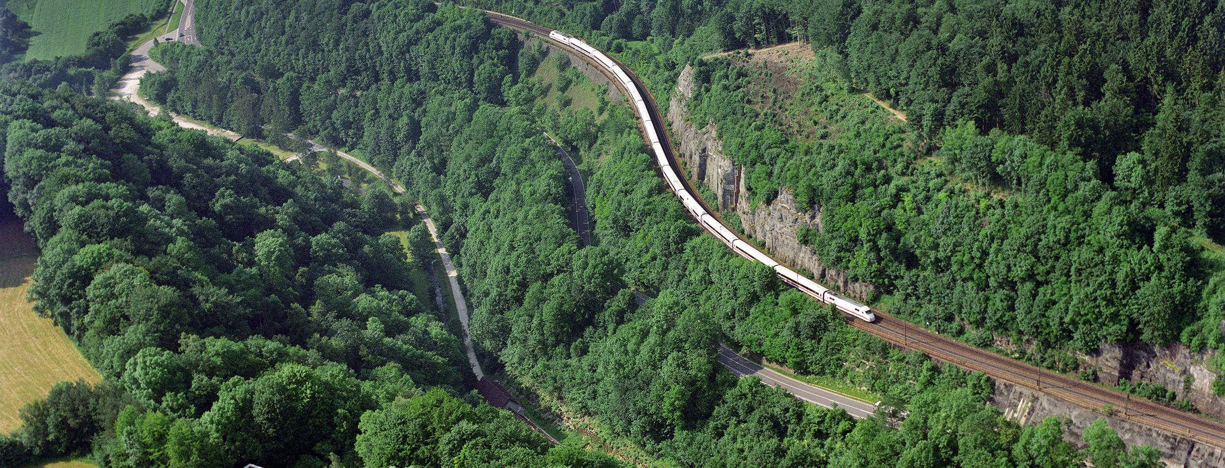 Luftbild zeigt einen weißen ICE-Zug wie er die Schienenstrecke an einem Berg hochfährt.