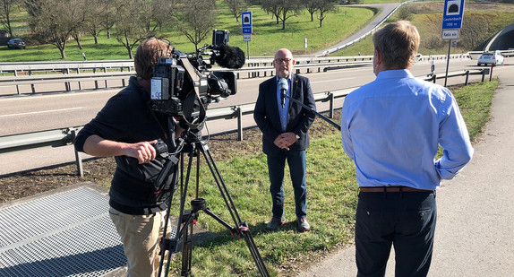 Verkehrsminister Winfried Hermann an einem Straßentunnel und wird von einem Fernsehereporter zusammen mit Kameramann interviewt. 