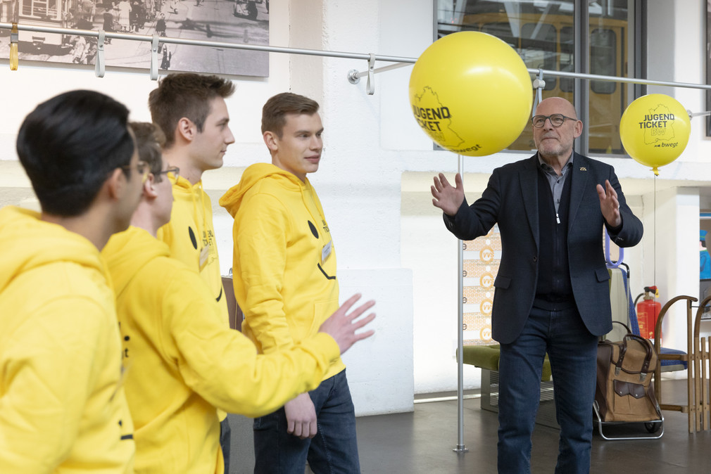 Minister Hermann und vier junge Männer in gelben Hoodies werfen sich gelbe Luftballons mit JugendticketNW-Logo zu.