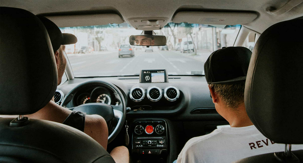 Ein Foto aus dem Innenraum eines Autos. Fotografiert von der Rückbank blickt man über die Schultern von zwei jungen Männer auf dem Fahrer- und Beifahrersitz.