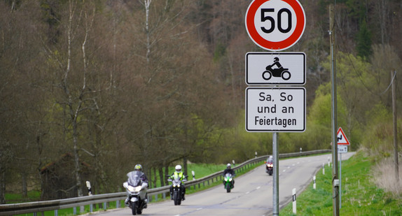 Ein Schild schreibt eine Geschwindigkeitsbeschränkung von 50 Kilometern pro Stunde für Motorräder an Samstagen, Sonntagen und Feiertagen vor. Im Hintegrund sind mehrere Motorradfahrerinnen und Motorradfahrer zu sehen.