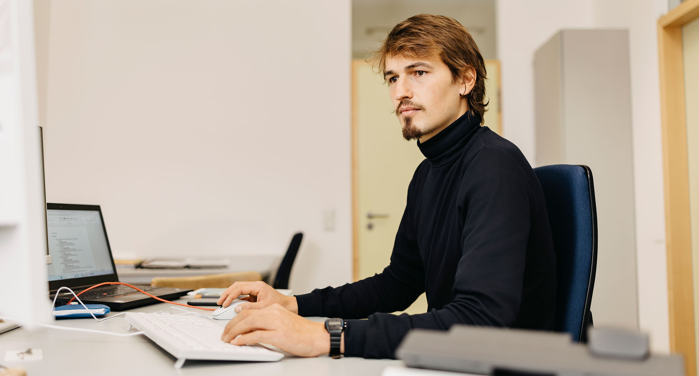 Ein junger Mann sitzt an einem Schreibtisch vor Bildschirmen und arbeitet.