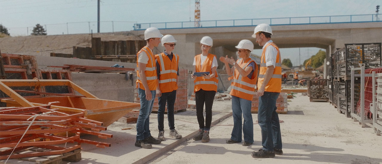 Auf einer Baustelle steht eine Gruppe von Frauen und Männern mit Helm und Sicherheitskleidung zusammen. Sie diskutieren.